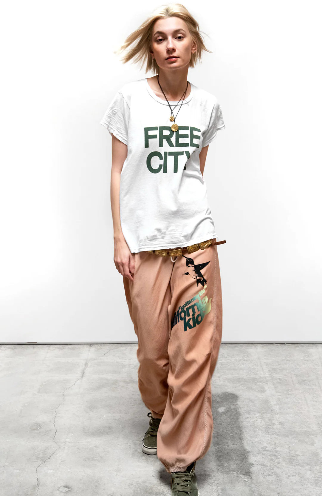 A person with short blonde hair wears a white "FREE CITY" t-shirt, Free City Loose Drawcord Biggy Pant, green sneakers, and layered necklaces, standing on a gray concrete floor against a plain white wall.