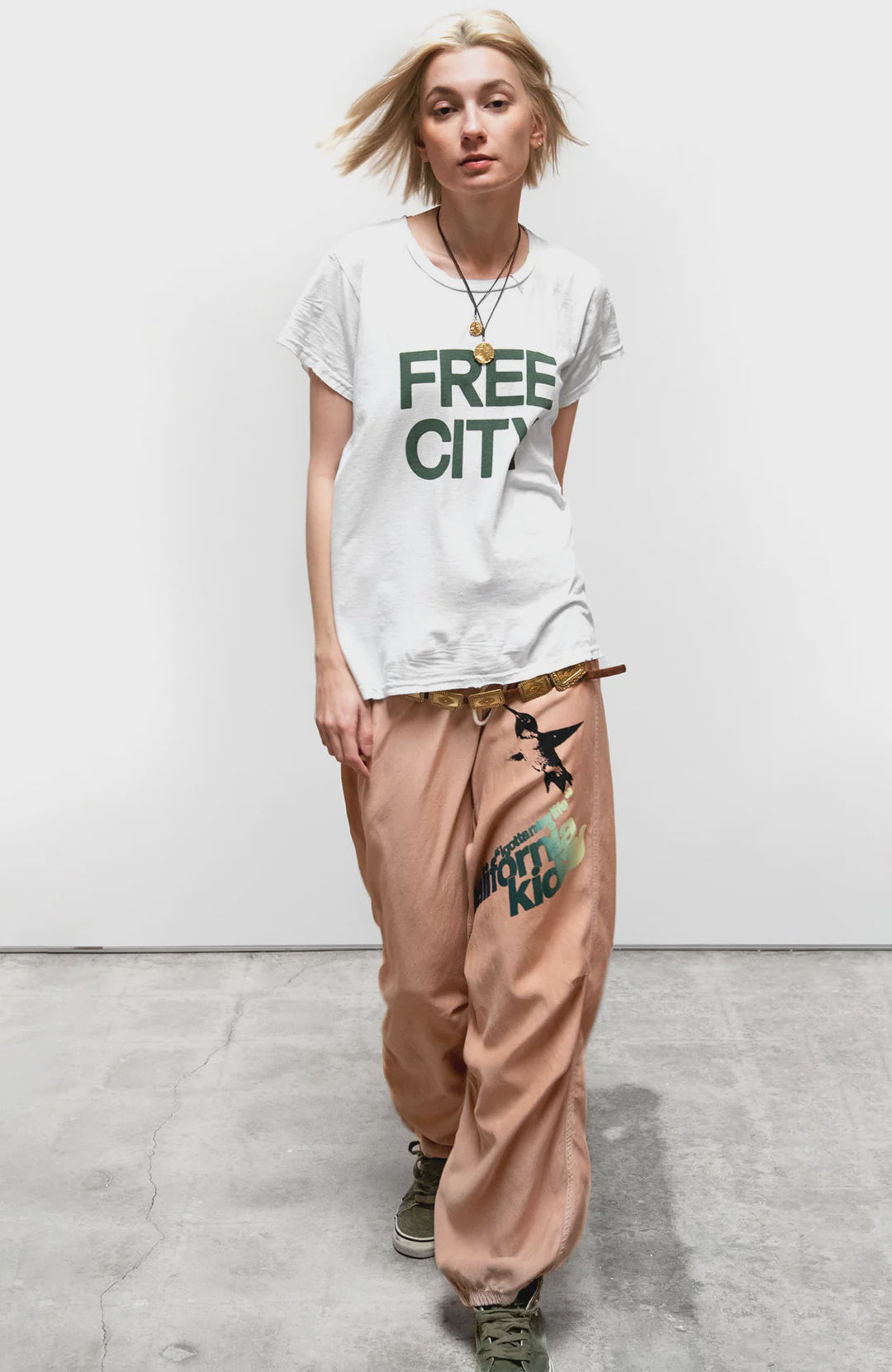 A blonde woman wears the FREECITY thedoingofitisthebeingofit unisex shortsleeve t-shirt by Free City (sparrow, LLC), loose tan pants with black print, green sneakers, and necklaces, standing against a white wall on a concrete floor.