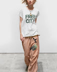 A blonde woman wears the FREECITY thedoingofitisthebeingofit unisex shortsleeve t-shirt by Free City (sparrow, LLC), loose tan pants with black print, green sneakers, and necklaces, standing against a white wall on a concrete floor.