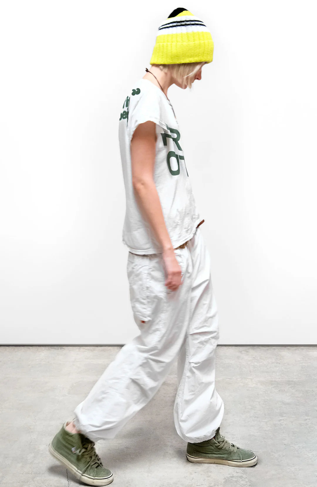 A person in a yellow-and-white beanie, white FREECITY thedoingofitisthebeingofit shortsleeve tee by Free City (sparrow, LLC), hand-screen printed in Los Angeles, white baggy pants, and green sneakers walks sideways on a gray floor.
