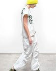 A person in a yellow-and-white beanie, white FREECITY thedoingofitisthebeingofit shortsleeve tee by Free City (sparrow, LLC), hand-screen printed in Los Angeles, white baggy pants, and green sneakers walks sideways on a gray floor.