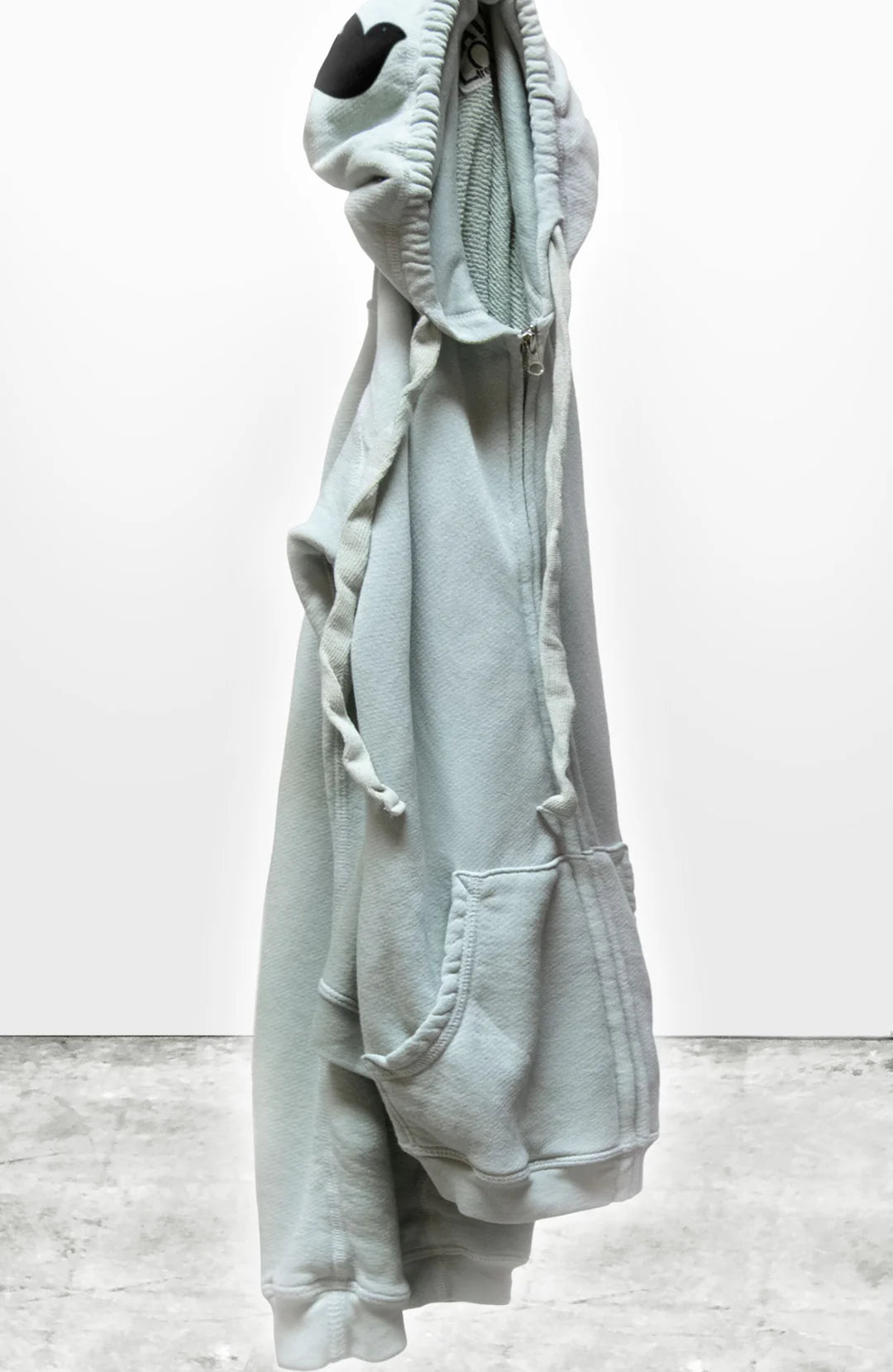 A light gray Free City SUPERFLUFF Lux Zip Hoodie by sparrow, LLC is hanging suspended by its hood against a plain backdrop. The front zipper is partially visible, with two front pockets and loosely hanging drawstrings. The concrete-like floor subtly peeks out at the bottom of this unisex hoodie.