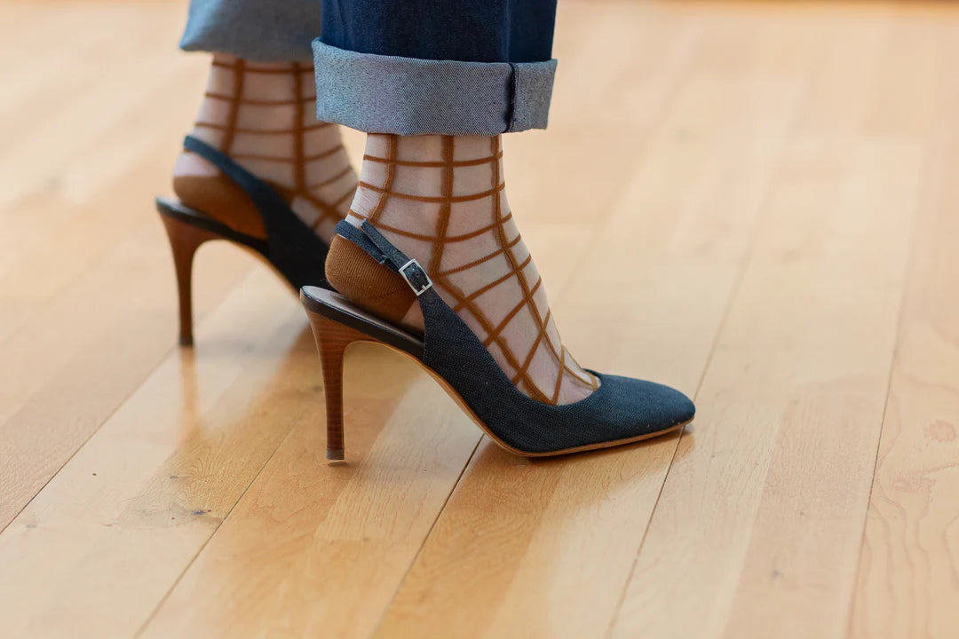 A person stands on a light wooden floor wearing rolled-up jeans, Unified Sheer French Window ankle socks, and navy blue high-heeled slingback shoes.