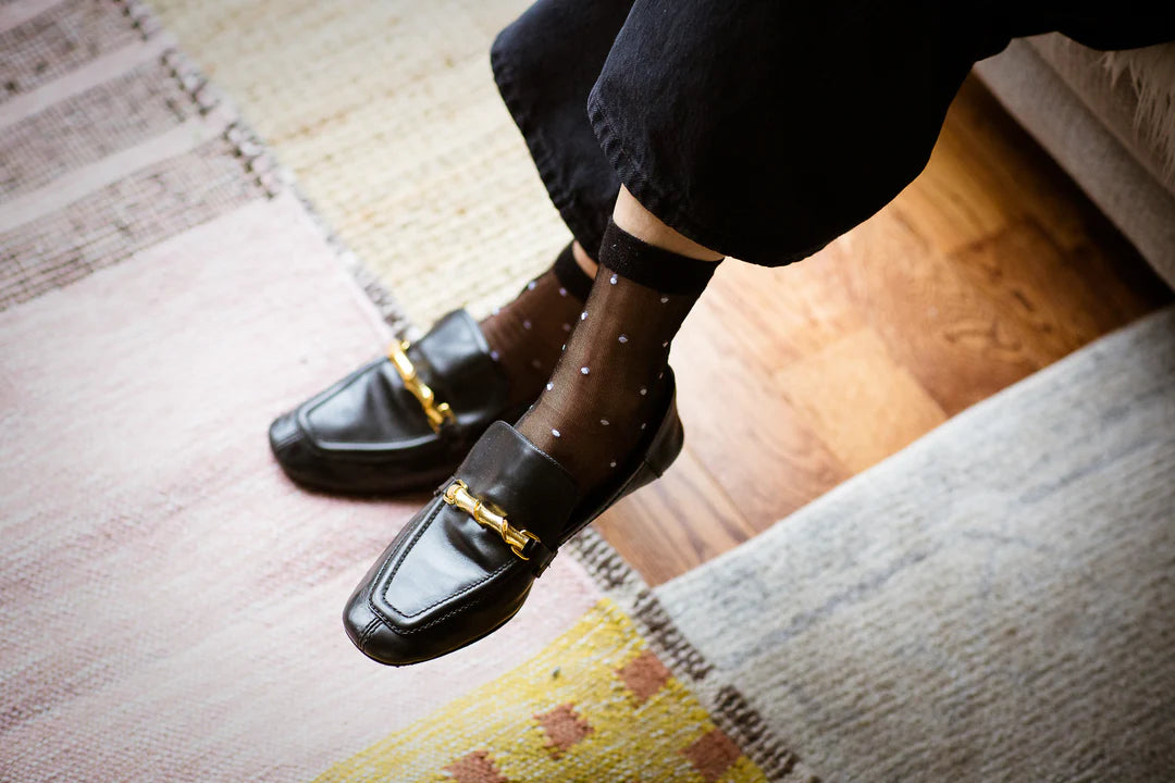 A person wearing Unified Sheer Dot Socks—stylish sheer black women's socks—and black loafers with gold hardware, sits cross-legged on a light-colored carpeted floor.