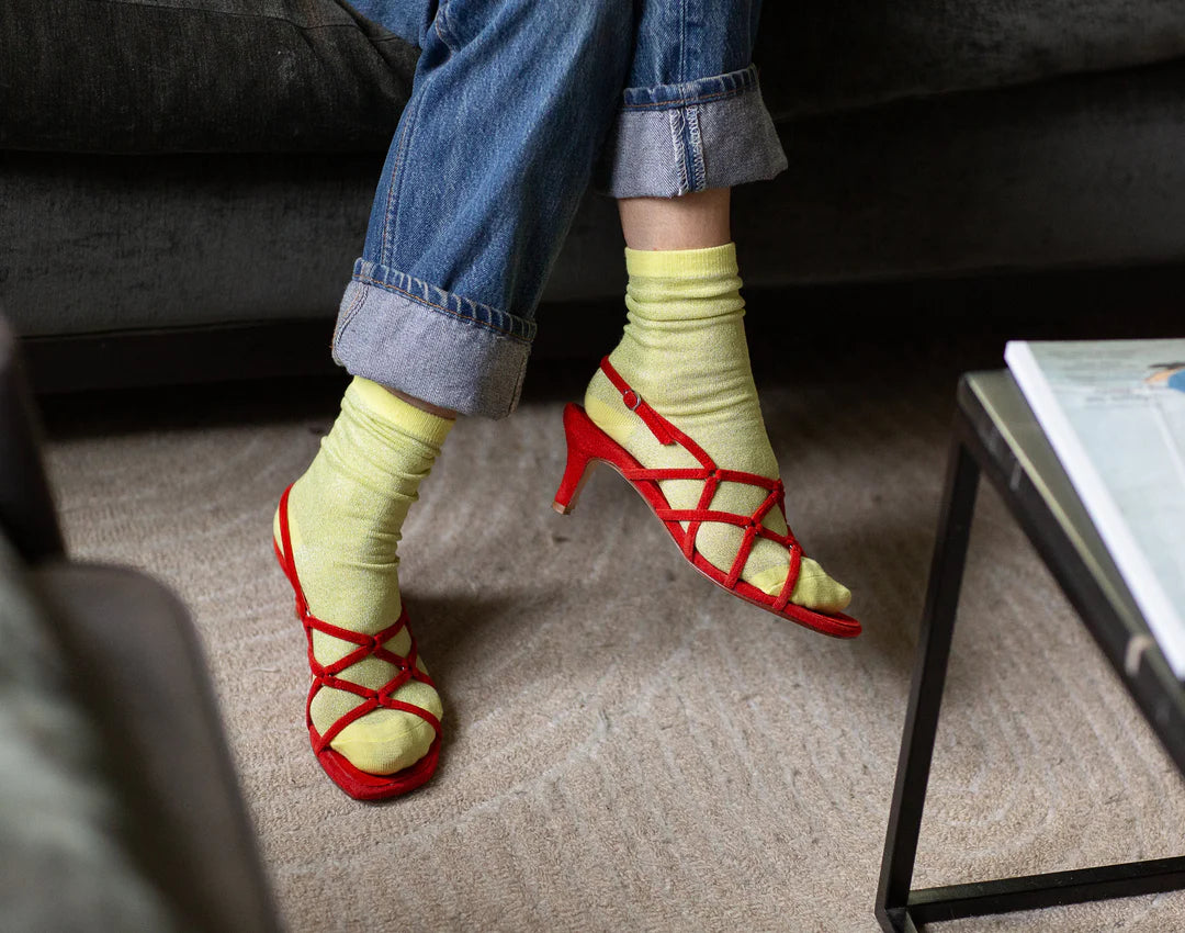 Someone sits cross-legged on a sofa, wearing Unified Glitter Socks with red strappy high-heeled sandals and rolled-up blue jeans, on a beige carpet beside a white coffee table.