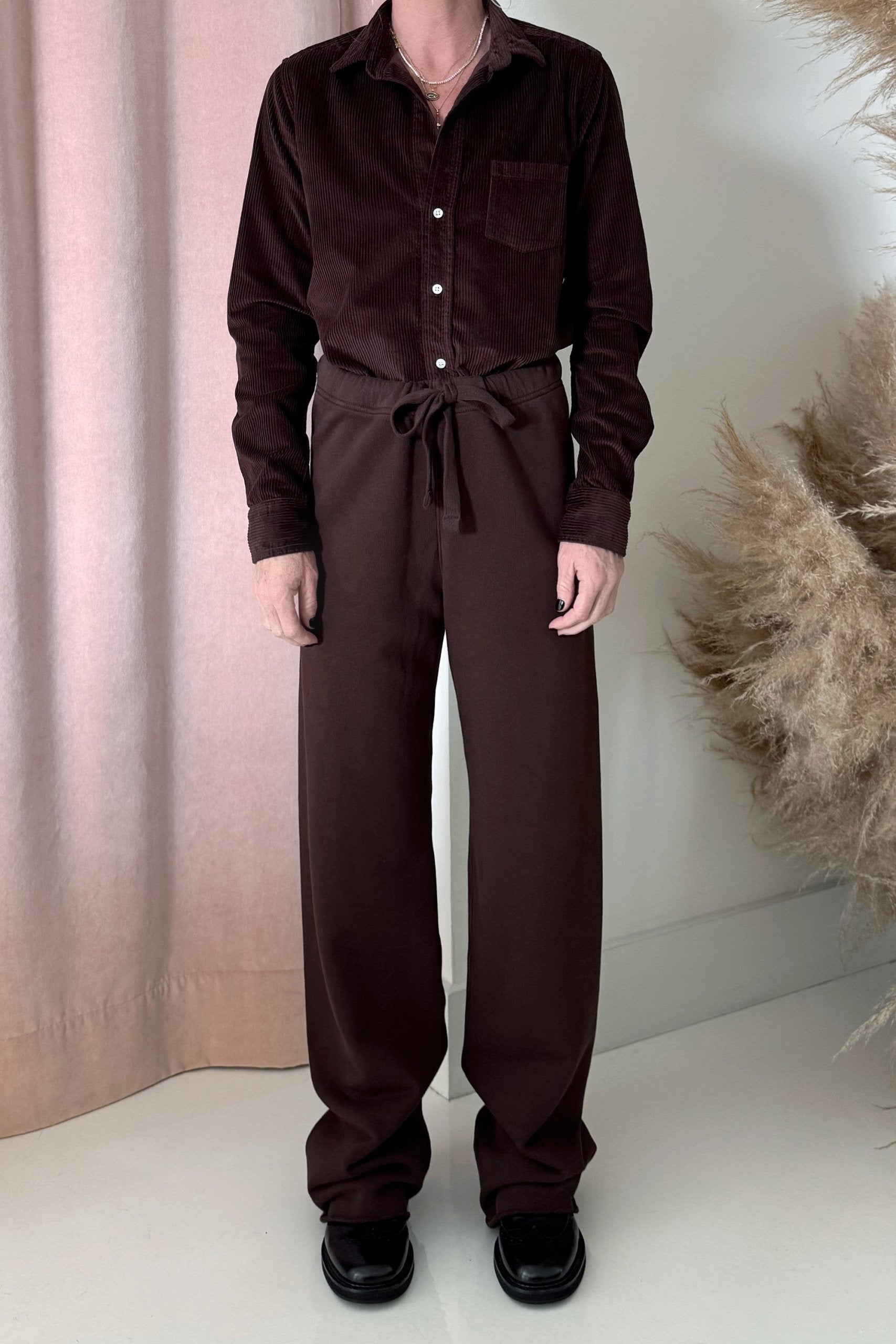 A person stands indoors in a dark brown button-up shirt and Frank & Eileen’s Catherine Long Sweatpant, made from 100% cotton with a waist tie. They wear black shoes, with pink curtains and beige pampas grass as background decor.