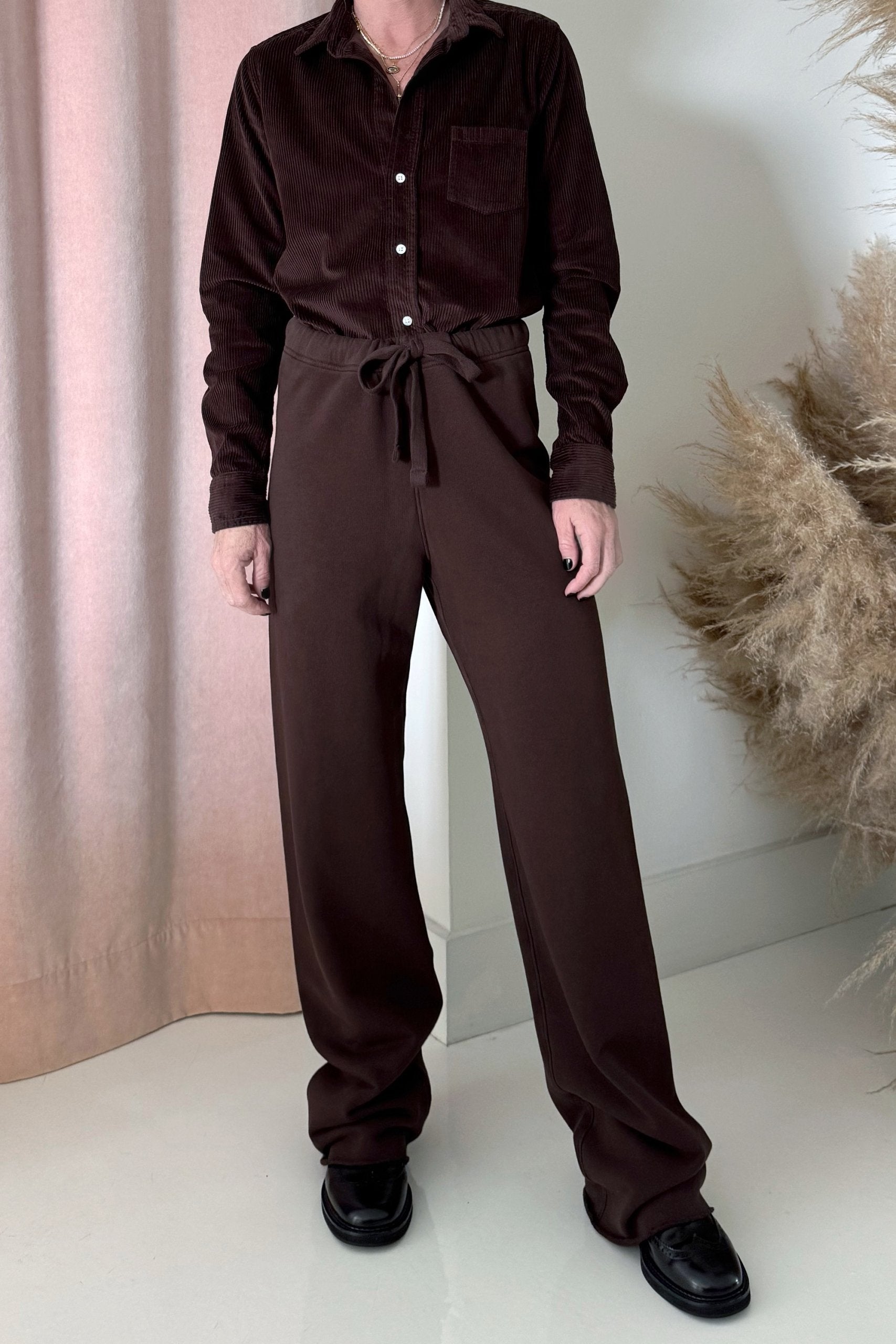 A person stands indoors wearing Frank & Eileen’s Catherine Long Sweatpant, paired with a dark brown corduroy button-up shirt and black shoes. Light pink curtains and dried grasses complete the background.