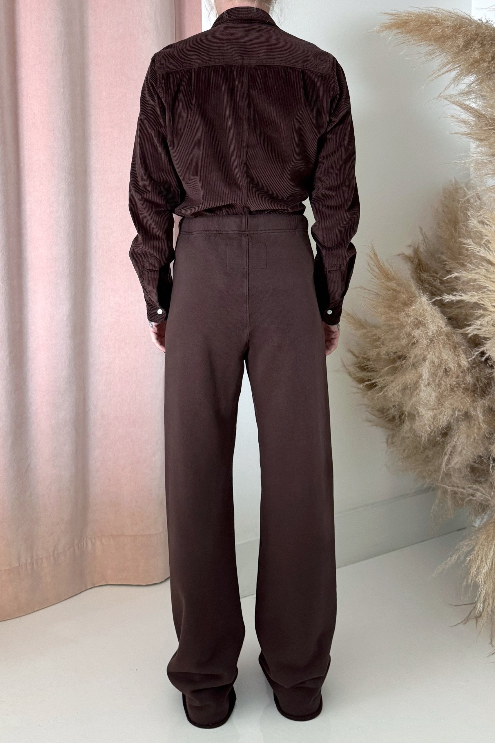 A person stands facing away, wearing Frank & Eileen’s Catherine Long Sweatpant with a dark brown long-sleeve shirt. Beige pampas grass and pink curtains create a soft backdrop.
