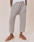 Against a cream fabric backdrop, someone wears The Great Inc.'s gray Jersey Crop harem pants with a dropped rise and brown moccasin-style shoes. The loose-fit design, similar to wider straight leg pants, pairs hands in pockets with a relaxed white top.
