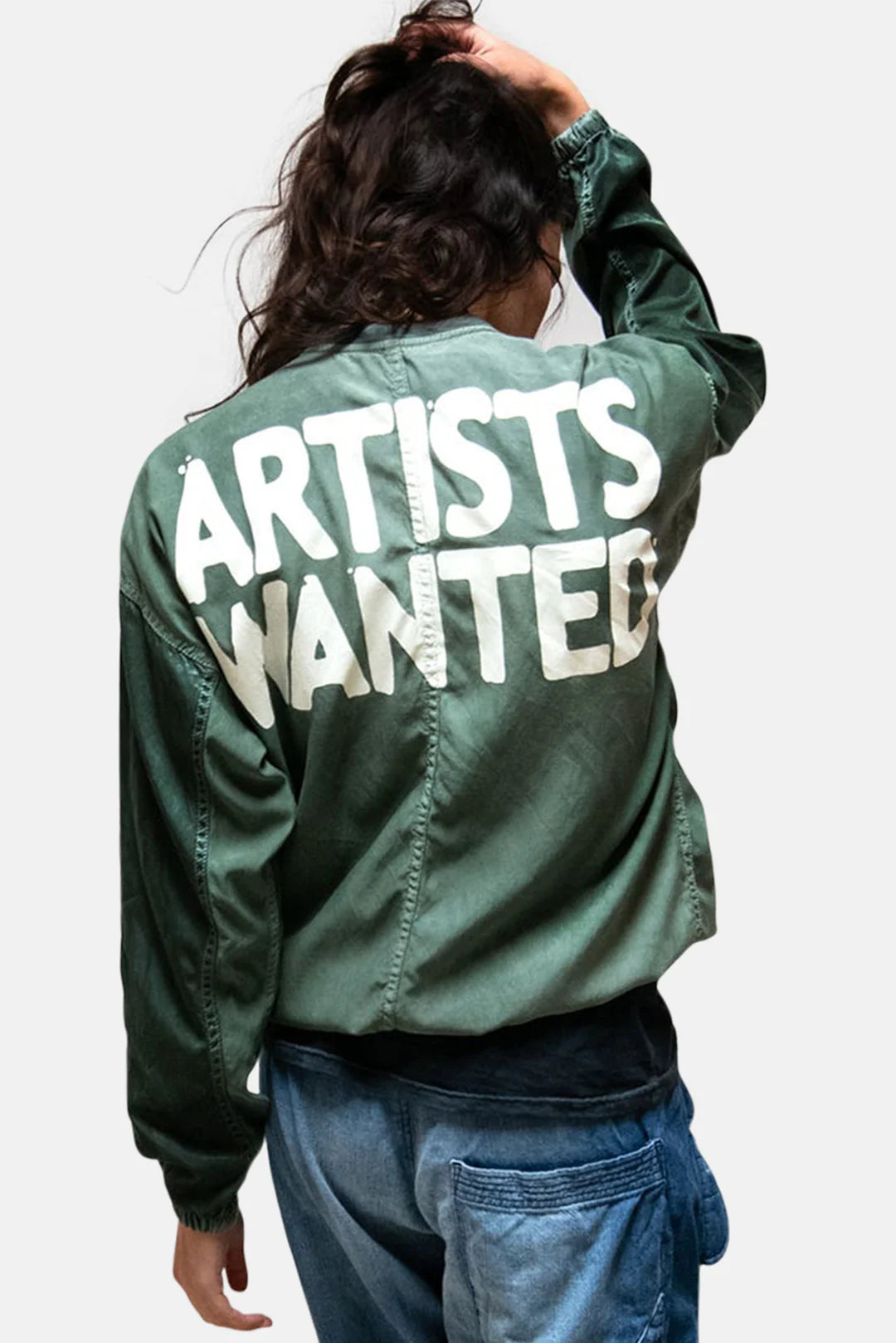 Seen from behind, a person with long dark hair wears the Free City (sparrow, LLC) Artist Wanted Matte Satin Air Jump Shell, featuring "ARTISTS WANTED" in bold white letters on the back, paired with blue jeans.