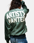 Seen from behind, a person with long dark hair wears the Free City (sparrow, LLC) Artist Wanted Matte Satin Air Jump Shell, featuring "ARTISTS WANTED" in bold white letters on the back, paired with blue jeans.