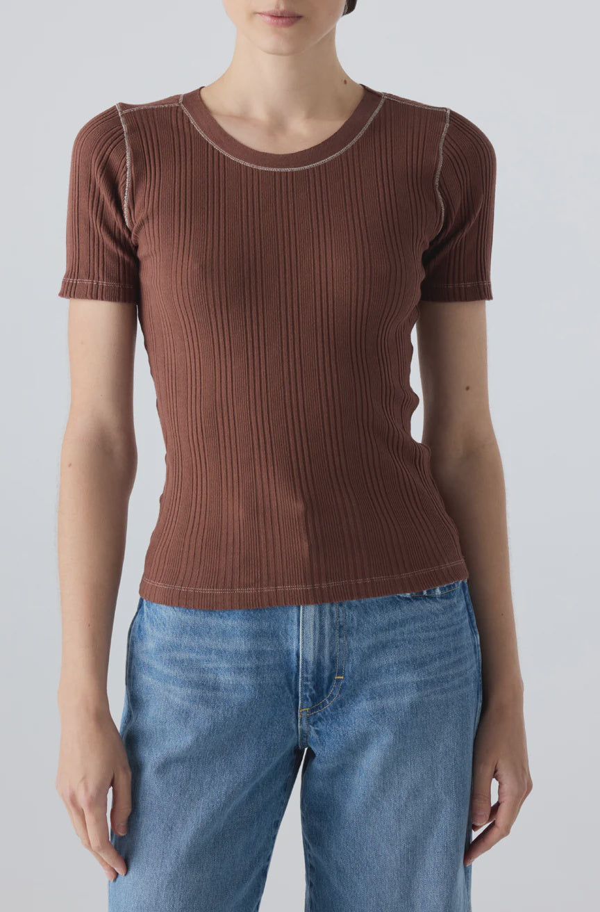 A person wearing the Cherish Tee by AMO Denim—a fitted, ribbed cotton tee with short sleeves and white seam stitching—paired with blue jeans, stands against a plain light gray background.