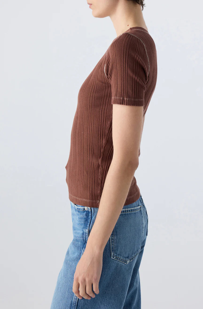 Wearing the AMO Denim Cherish Tee, a ribbed cotton top with a fitted silhouette, paired with light blue jeans and standing in profile against a plain light background.
