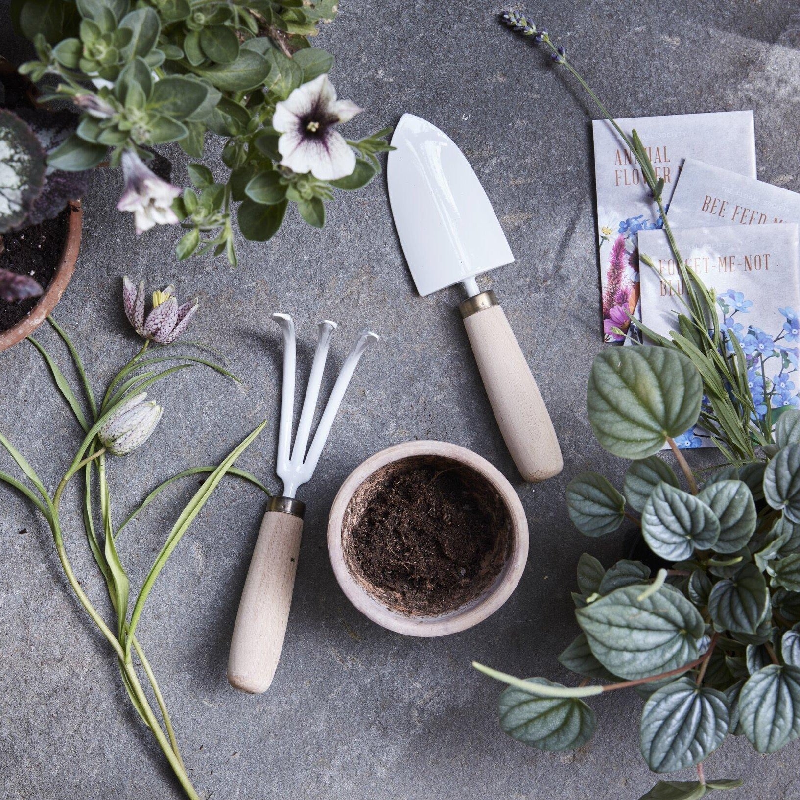 A serene gardening setup features the Garden Tool Set by The Floral Society, including a hand-crafted garden trowel made with Japanese stainless steel and a hand rake with wooden handles, lying on a stone surface. They are surrounded by potted plants, soil in a bowl, and seed packets.