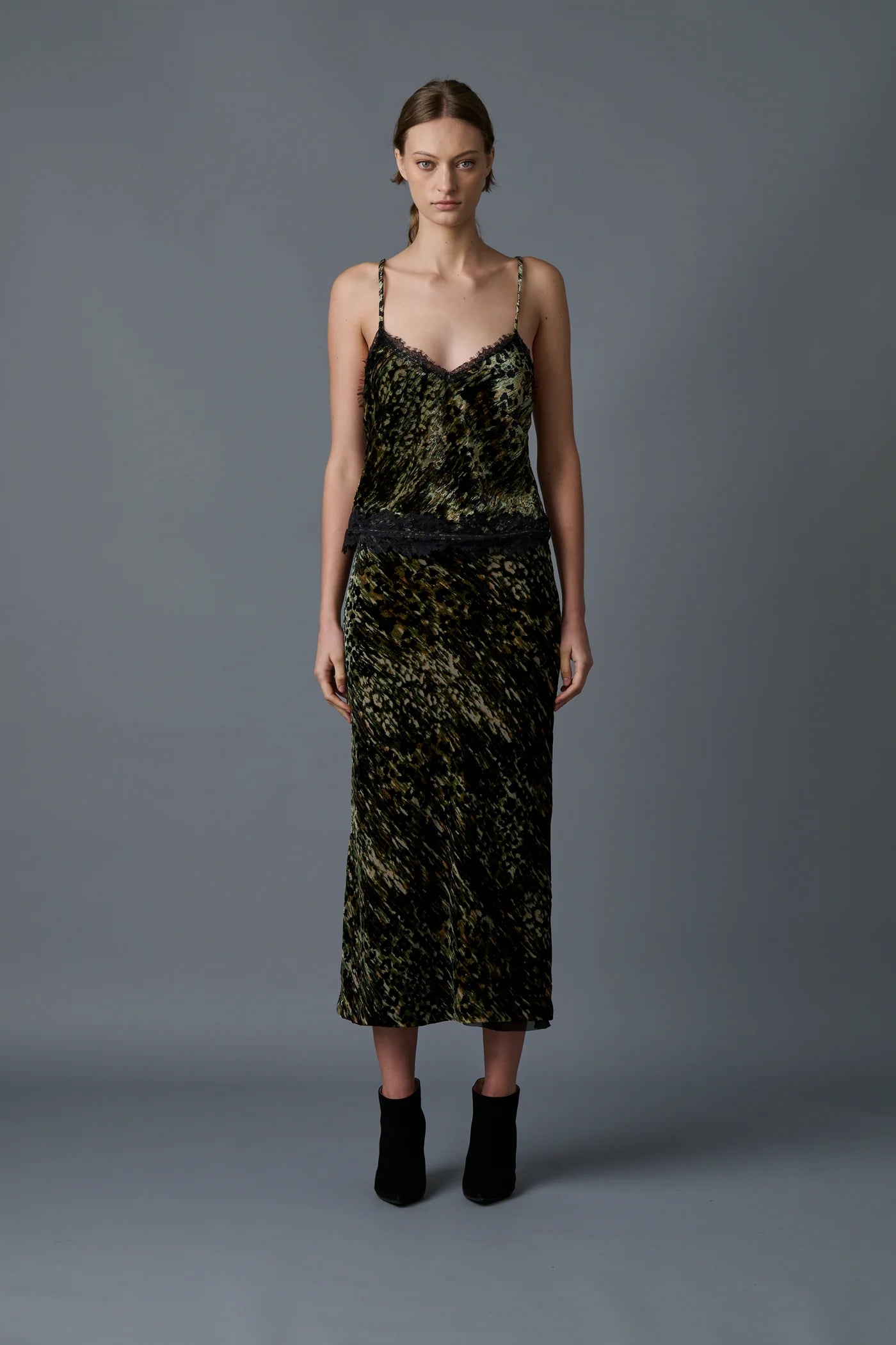 A woman stands against a grey background wearing the Velvet Underground Skirt by Le Superbe, featuring an abstract cheetah print, thin straps, and lace detail at the waist. She has shoulder-length hair styled simply and wears black ankle boots. She gazes directly at the camera with a neutral expression.