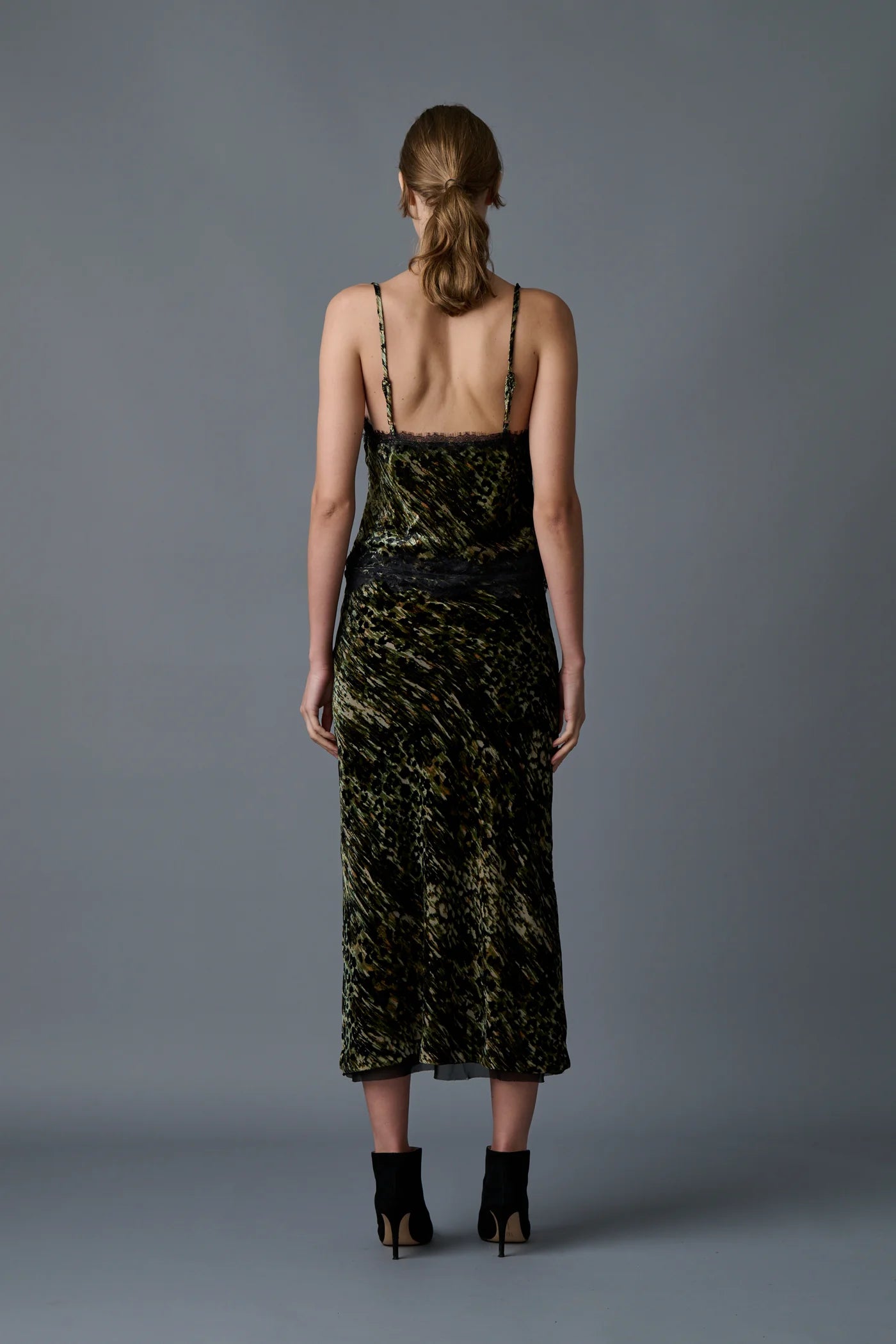 A person with light brown hair styled in a low ponytail is standing with their back to the camera. They are wearing the Velvet Underground Skirt by Le Superbe, which features a strappy, abstract cheetah print design, and black high-heeled sandals. The background is a plain, dark gray.