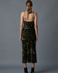 A person with light brown hair styled in a low ponytail is standing with their back to the camera. They are wearing the Velvet Underground Skirt by Le Superbe, which features a strappy, abstract cheetah print design, and black high-heeled sandals. The background is a plain, dark gray.