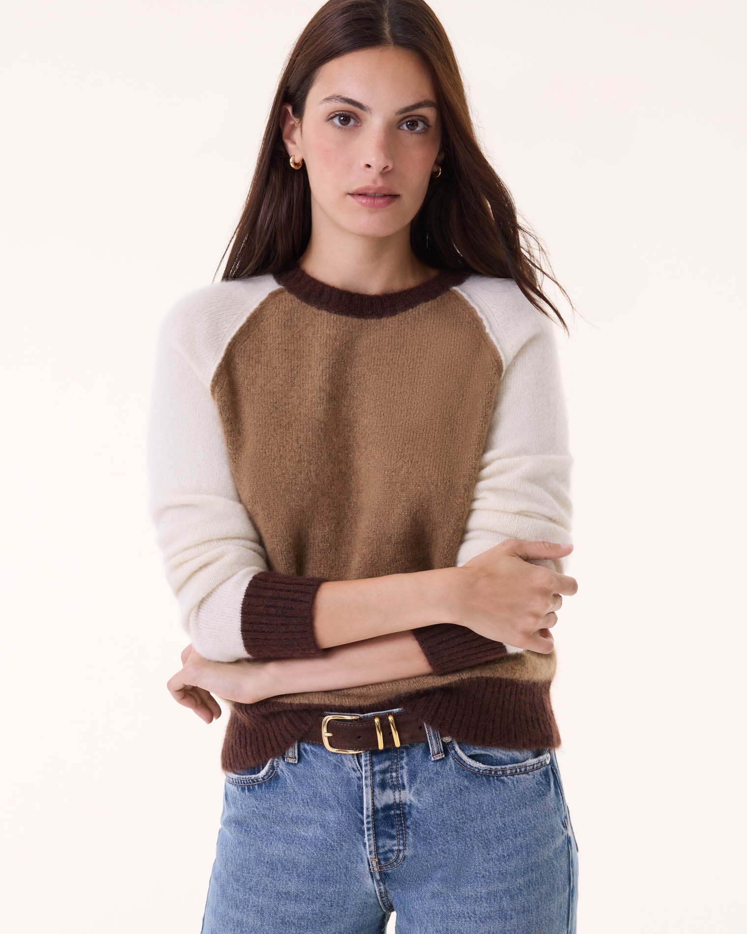 A woman with long brown hair models the BCS Group Nimbus Sweater, a color-block round neck style in brown, tan, and cream, paired with blue jeans and a black belt against a plain light background.