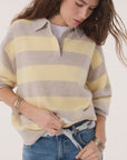 Wearing the KUJTEN Royde Stripe Sweater in yellow and beige over a white shirt with blue jeans and silver bangles, a person looks down while tucking in their shirt.