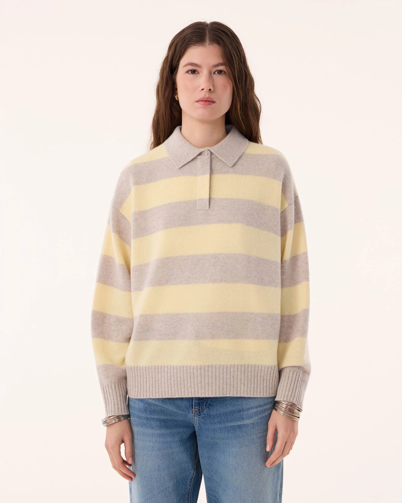 A woman with long brown hair wears the KUJTEN Royde Stripe Sweater—a yellow and gray collared rugby top—paired with blue jeans, standing against a plain light background.