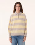 A woman with long brown hair wears the KUJTEN Royde Stripe Sweater—a yellow and gray collared rugby top—paired with blue jeans, standing against a plain light background.