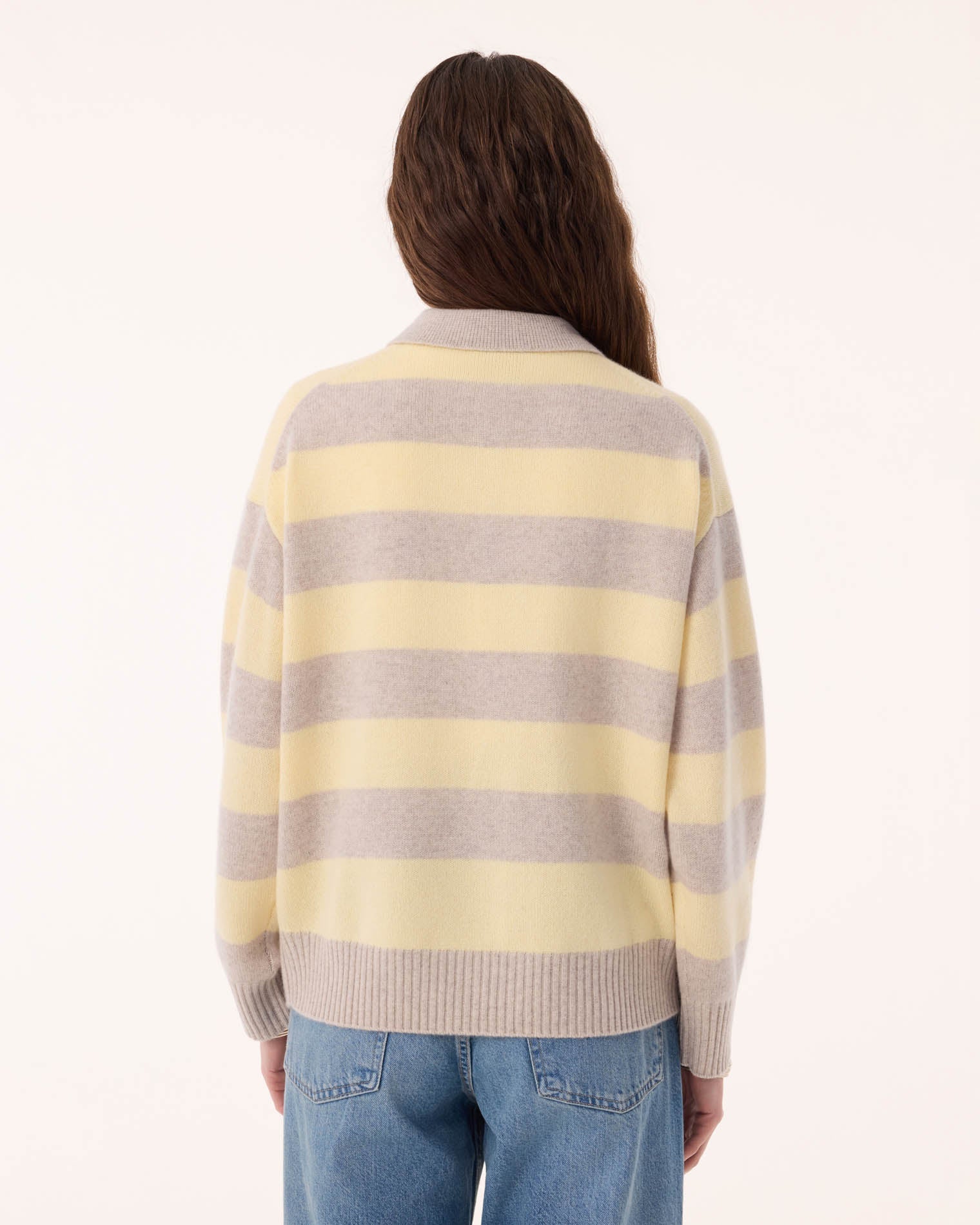 A person with long brown hair, seen from behind, wears the KUJTEN Royde Stripe Sweater with blue jeans against a plain light background.
