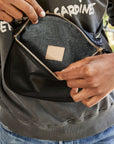 Close-up of hands opening the Clare Vivier Fanny Pack Fall 25, crafted in black natural grain leather with a gold zipper. Fabric interior displays the "Clare V." label. The wearer, dressed in a dark sweatshirt, highlights this chic everyday accessory.