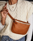 A person wears a cream cable-knit sweater, light-wash jeans, a gold necklace, and the Clare Vivier Fanny Pack Fall 25: a brown natural grain leather crossbody bag with zippered closure, worn hands-free across the chest.