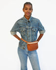 A woman in a denim jacket and jeans stands against a white background, hands on hips. She has braided hair and wears the Clare Vivier Fanny Pack Fall 25, crafted from natural grain leather, around her waist.