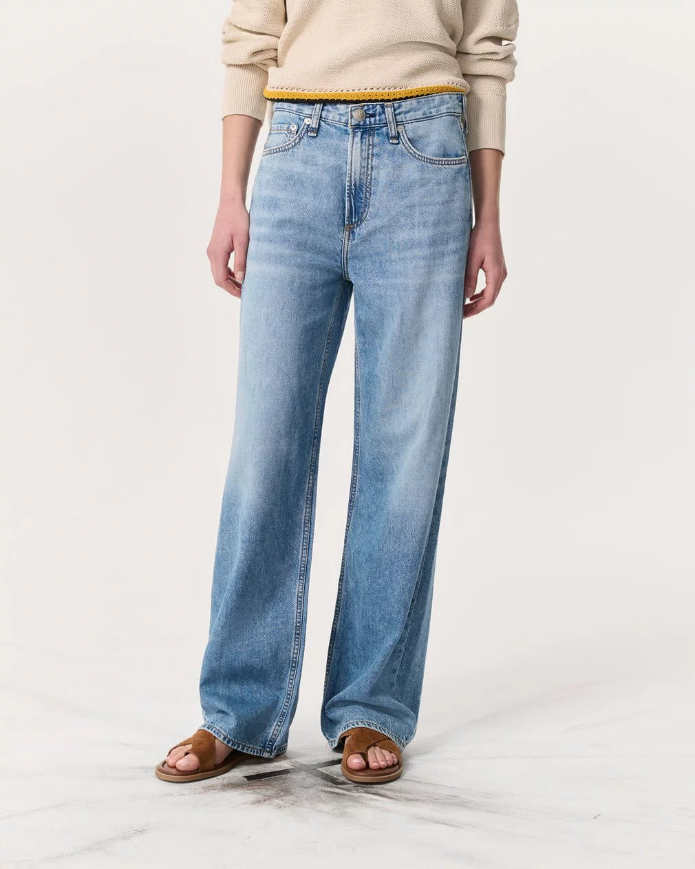 A person models Rag & Bone's Logan Pant in light blue, styled with a beige long-sleeve sweater, brown slide sandals, and a yellow belt against a plain white background. Their head is not visible in the image.