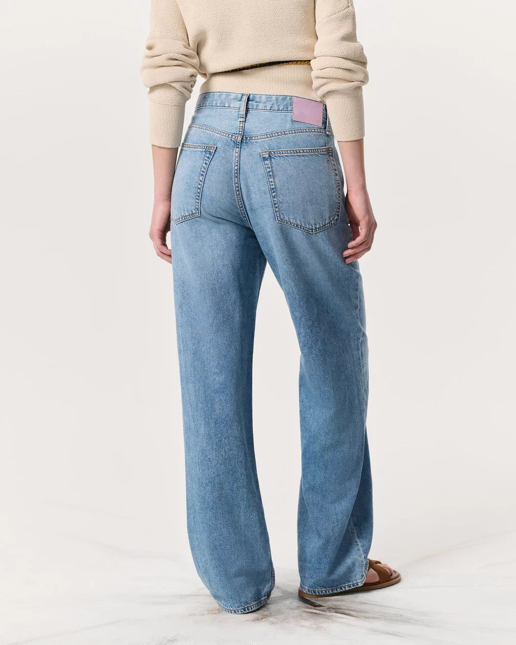A person wearing Rag & Bone's Logan Pant—light blue, high-waisted wide-leg jeans made from Featherweight denim—plus a beige sweater, seen from the back. The jeans have two back pockets and a pink label on the waistband.