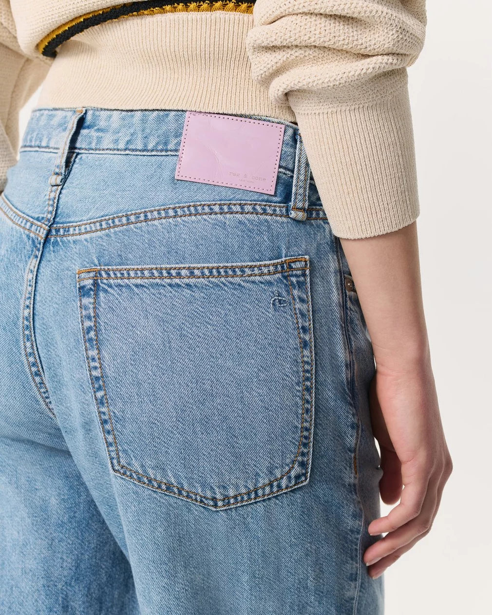 A person is shown from the back wearing Rag & Bone's Logan Pant in light blue Featherweight denim, paired with a cream knit sweater. The focus is on the jeans' back pocket and a pale pink leather patch on the waistband.