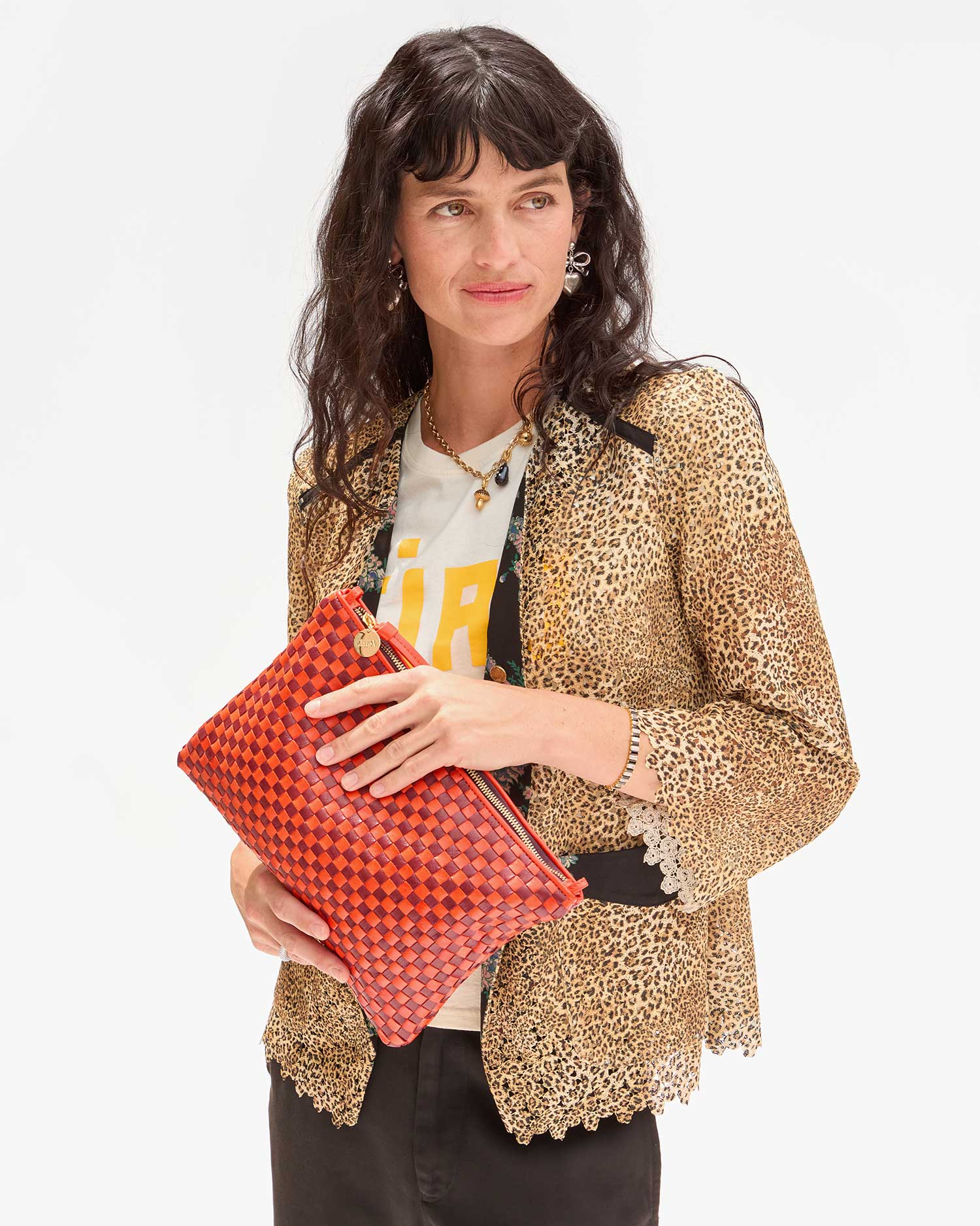 A person with long dark hair holds the vibrant Flat Clutch w/ Tabs by Clare Vivier. They wear a leopard print jacket over a t-shirt with yellow text and black pants, standing against a plain white background.