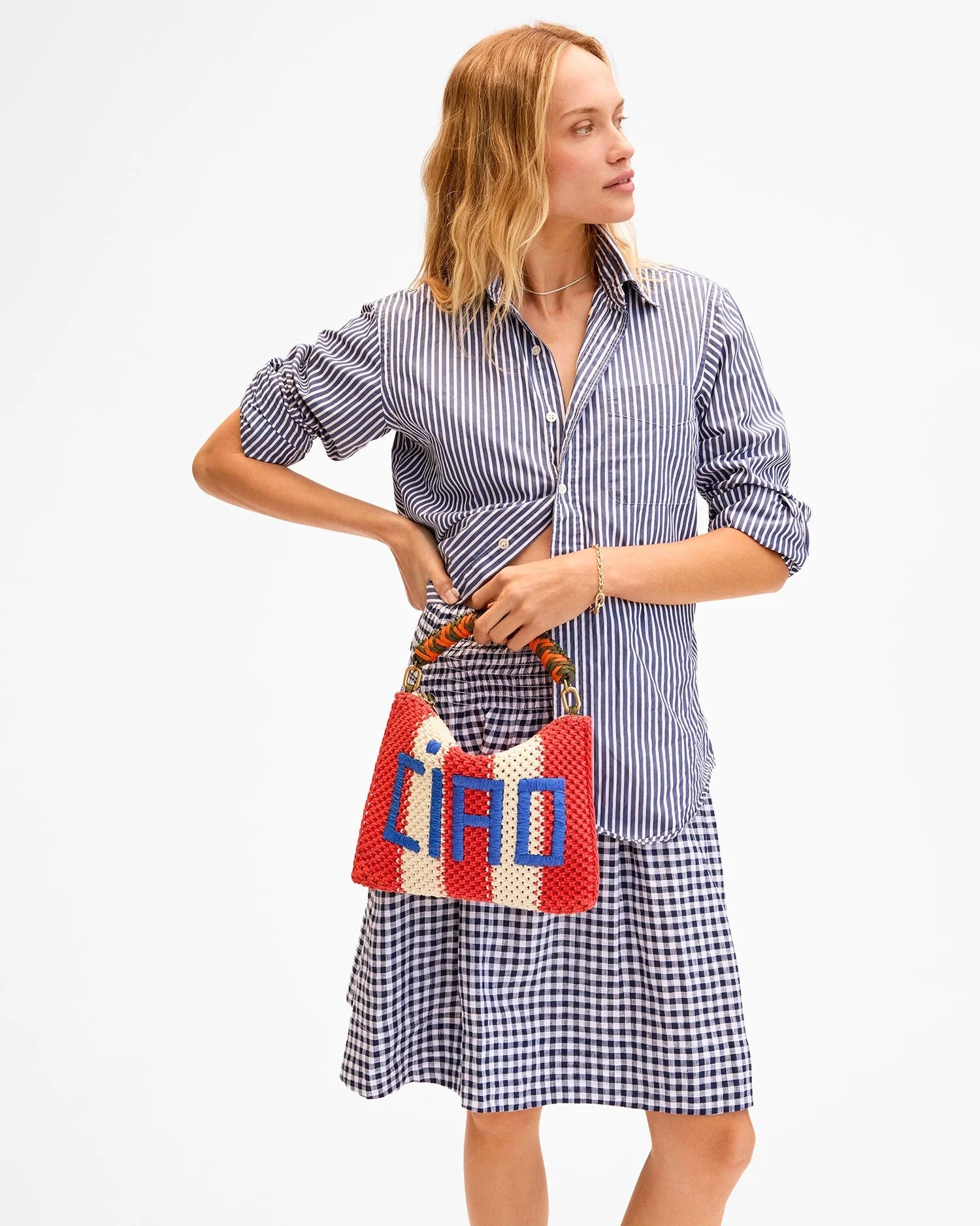 A woman in a blue and white striped button-down and gingham skirt holds the Clare Vivier Flat Clutch W/ Tabs S25, a red, cream, and blue crocheted cotton bag with bamboo handles, while looking to the side against a plain white background.