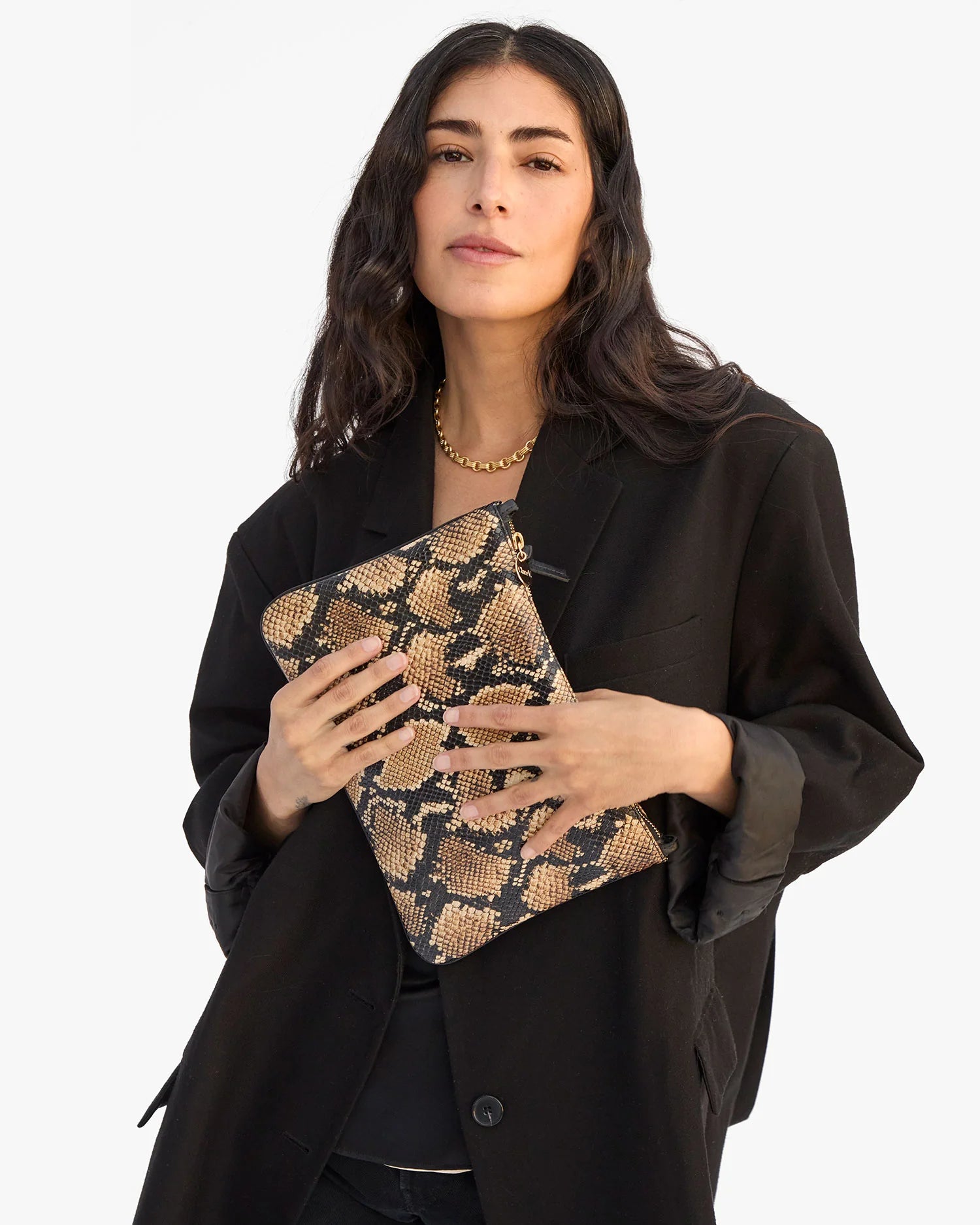 A woman with long dark hair wears a black blazer and holds the Clare Vivier Flat Clutch with Tabs 26, featuring a tan-and-black snakeskin pattern, as she poses against a plain light background.