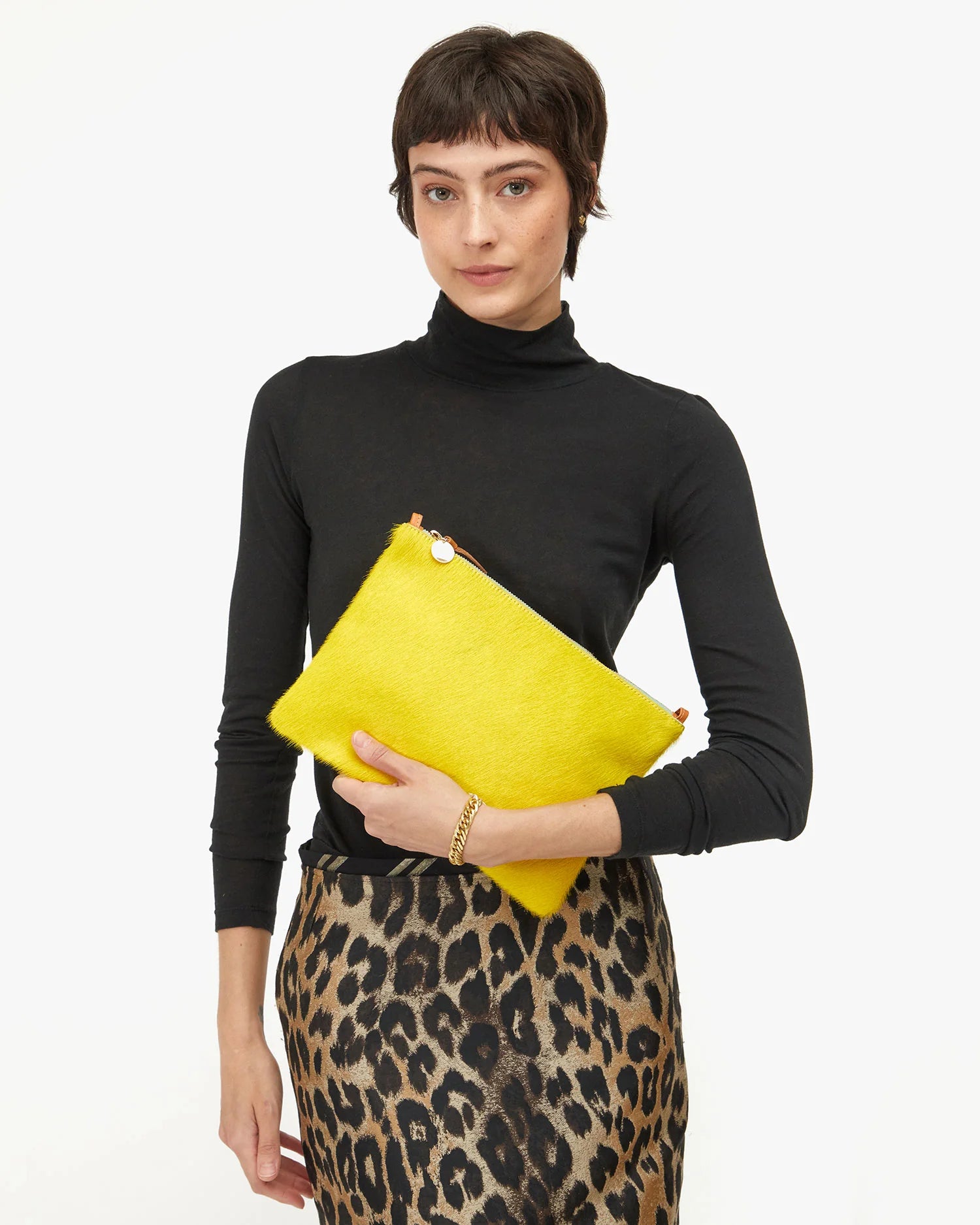 A woman with short brown hair in a black turtleneck and leopard print skirt holds the Clare Vivier Flat Clutch W/ Tabs S25, a bright yellow crocheted cotton bag, against a white background.