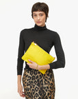 A woman with short brown hair in a black turtleneck and leopard print skirt holds the Clare Vivier Flat Clutch W/ Tabs S25, a bright yellow crocheted cotton bag, against a white background.