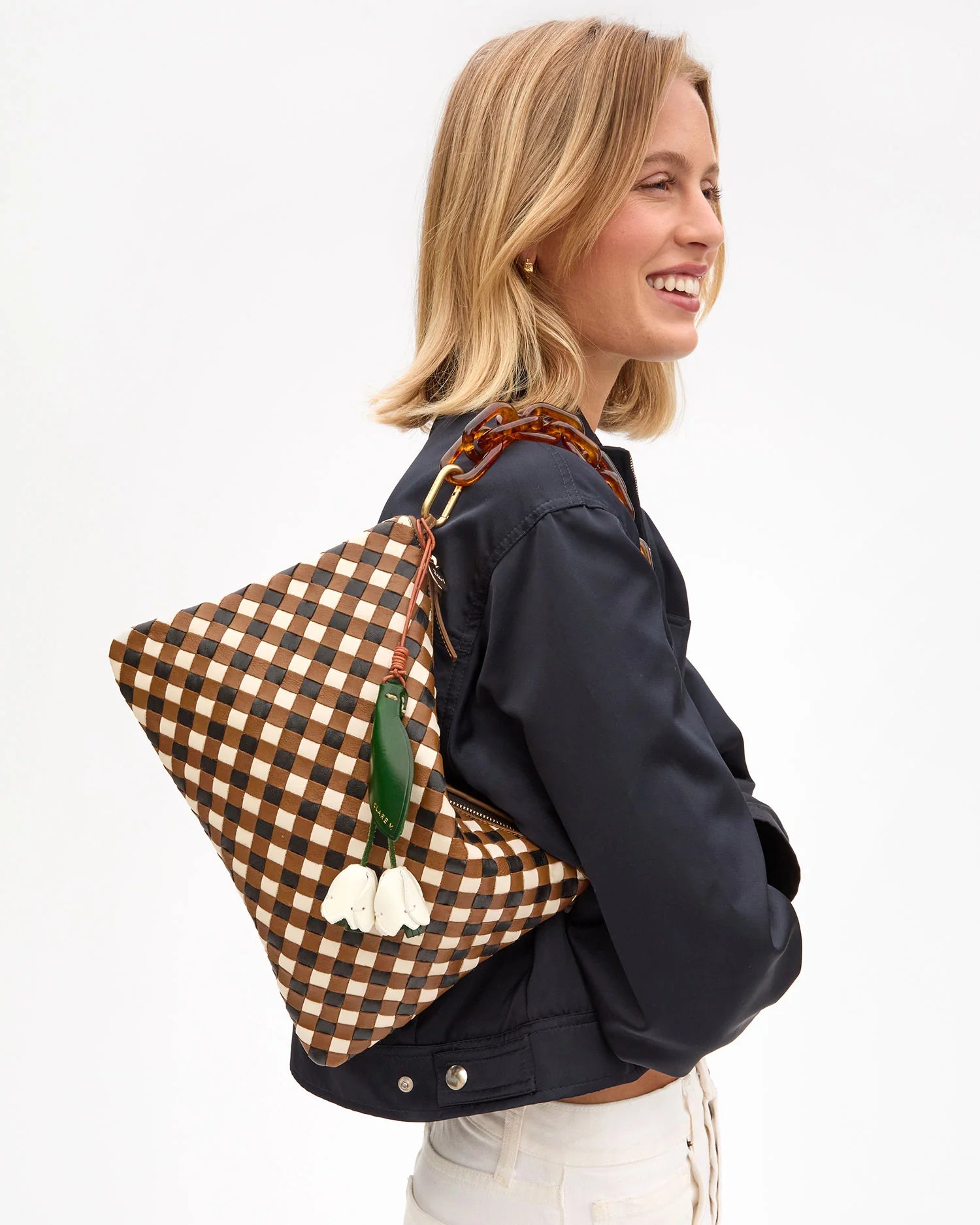 A smiling blonde woman wears a dark jacket and carries the Clare Vivier Flat Clutch with Tabs 26, featuring a handwoven leather design and a green and white flower charm on her shoulder.