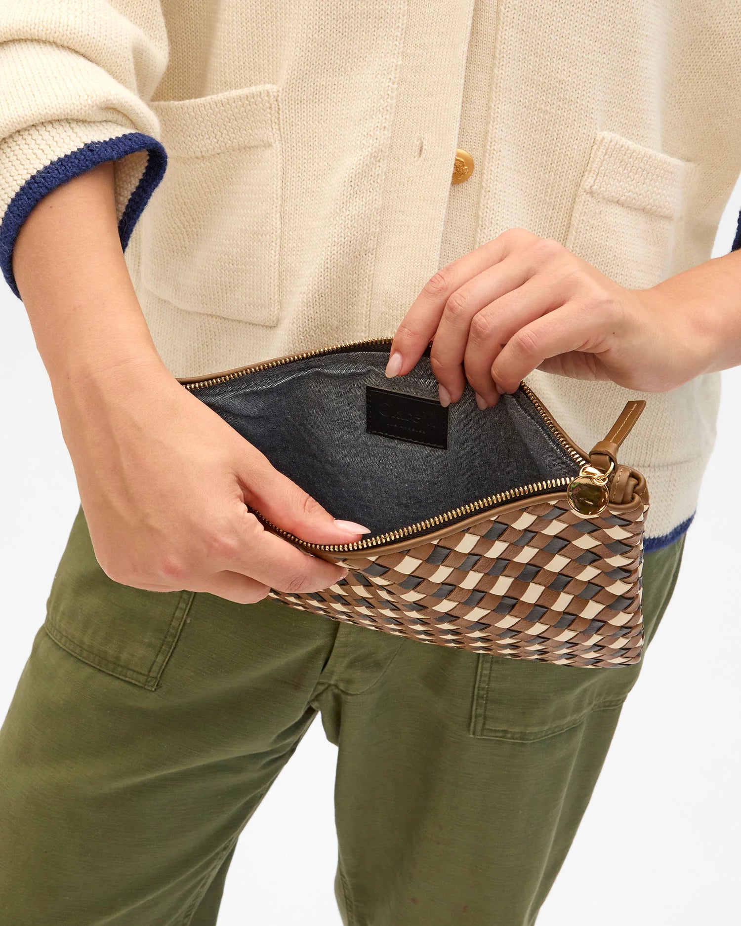 A person in a cream sweater and green pants holds the Clare Vivier Flat Clutch with Tabs 26, an open, handwoven leather zippered pouch, revealing its dark fabric interior.