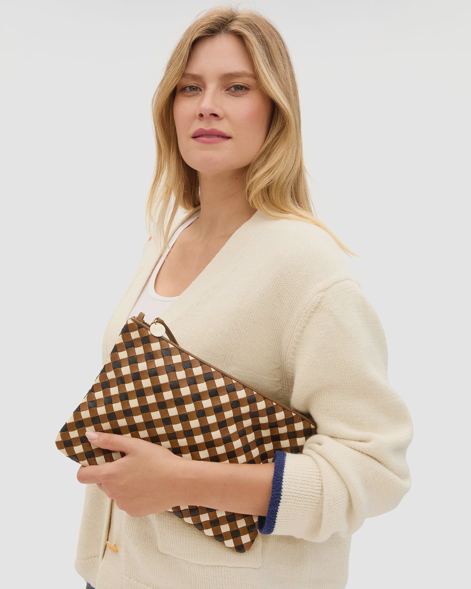 A woman with blonde hair, dressed in a cream cardigan, holds the Clare Vivier Flat Clutch with Tabs 26—a handwoven brown and black checked leather bag—against a plain, light background.