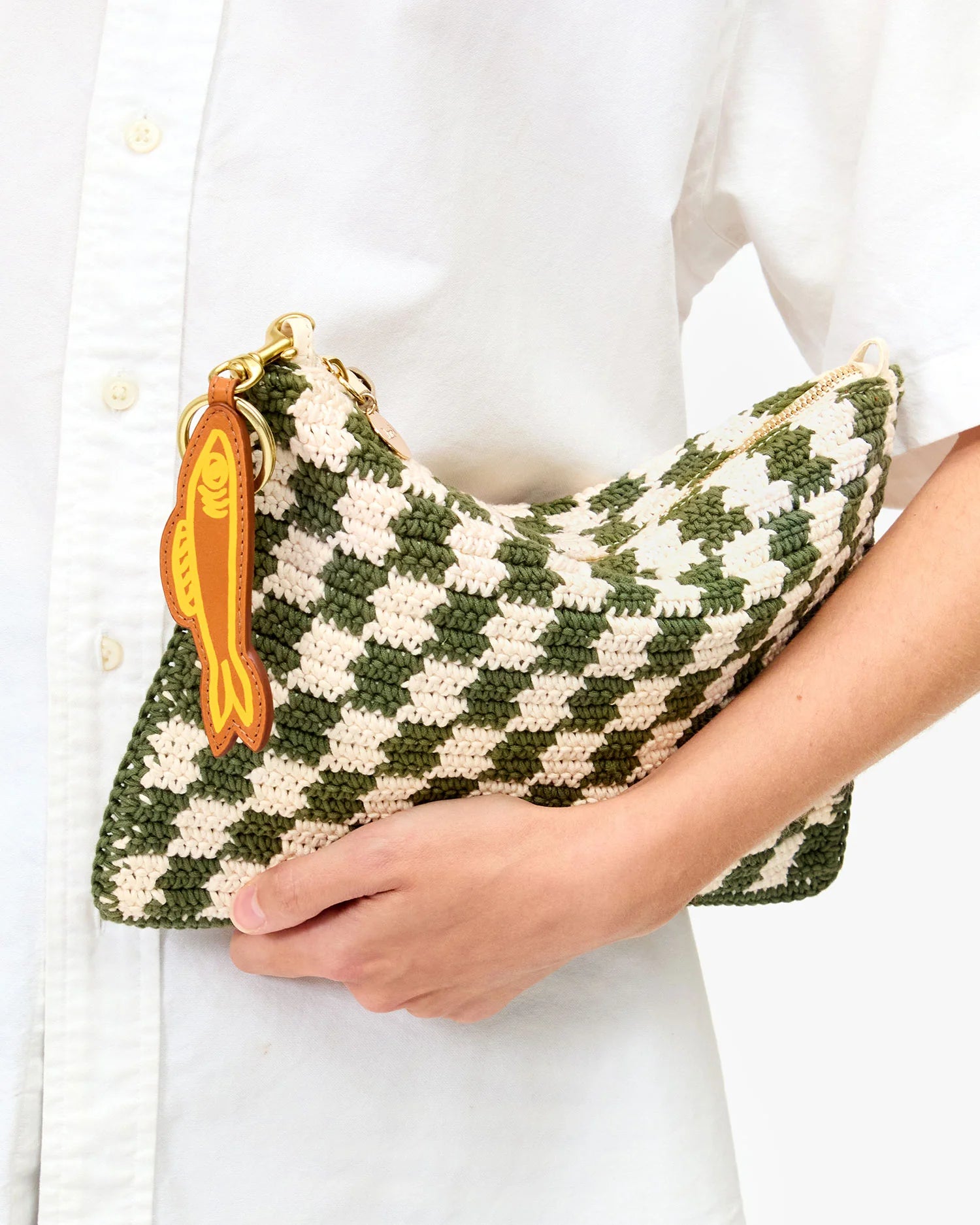 A person in a white button-up shirt holds the Clare Vivier Flat Clutch W/Tabs, a green and cream checkered cotton piece with a yellow fish-shaped keychain on the zipper.