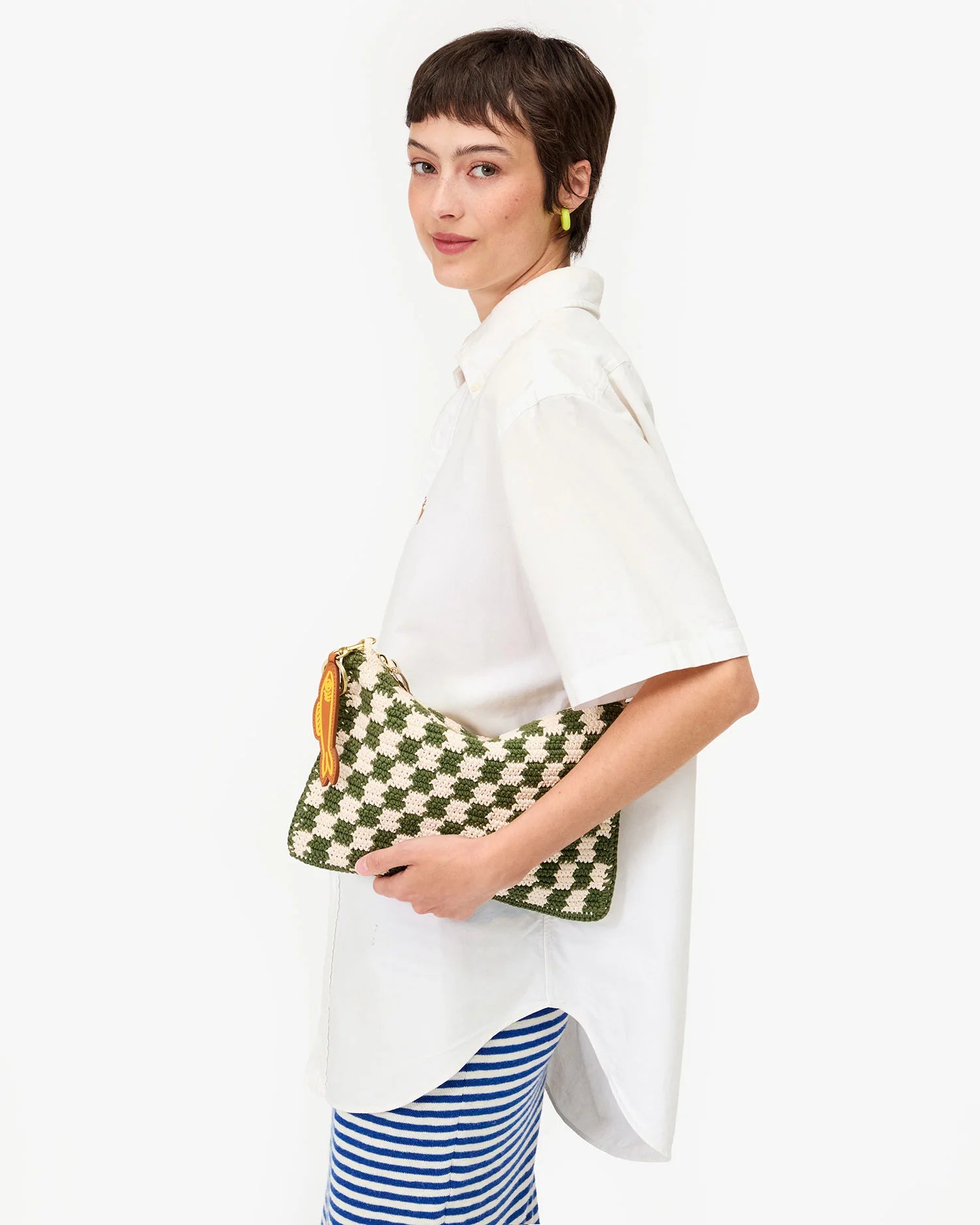 A woman with short brown hair holds a Clare Vivier Flat Clutch W/Tabs, a green and white crocheted cotton pouch with a yellow zipper, while wearing a loose white shirt over blue-and-white striped pants against a white background.