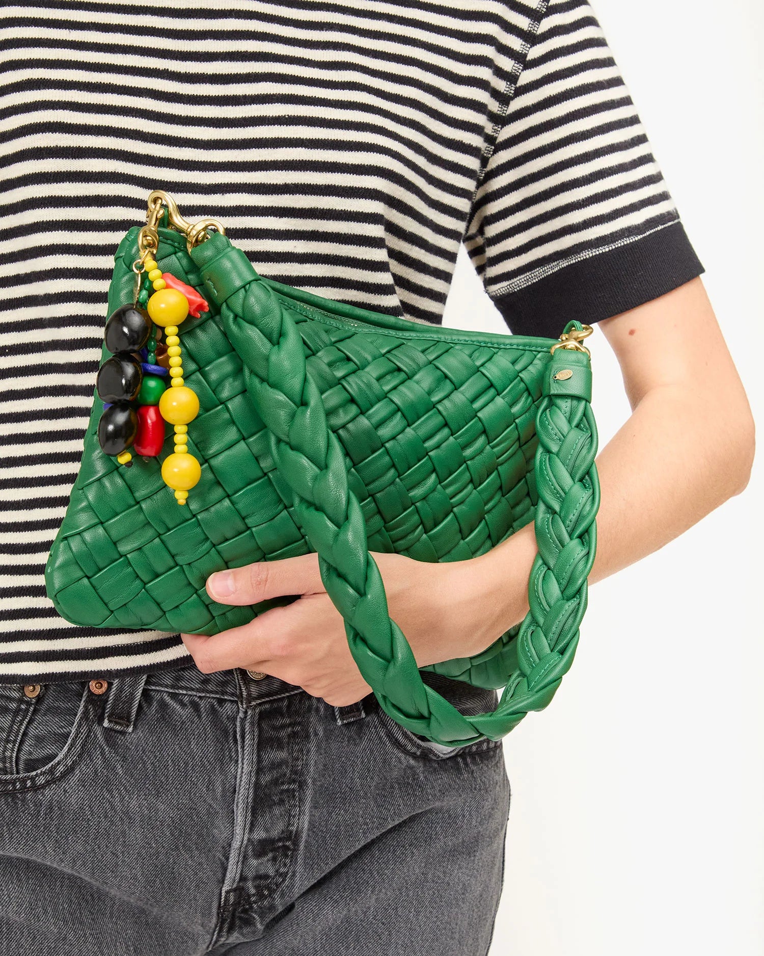 A person wearing a striped shirt and black jeans carries the Clare Vivier "Le Long Beaded Tassel," a green woven handbag with wood beads and colorful accents.