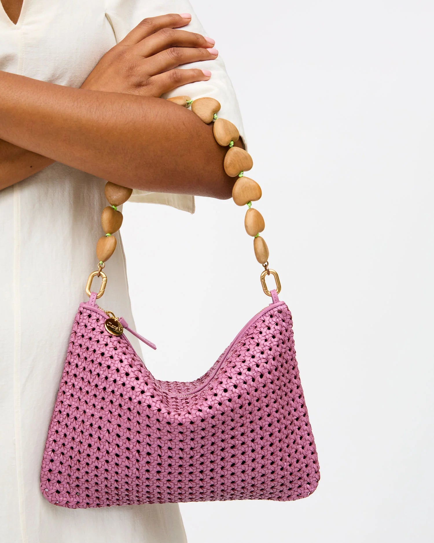 A person in a light-colored outfit holds Clare Vivier’s Shortie Strap/Heart, a pink woven handbag featuring a chunky strap made of large, irregular wooden beads, against a plain white background.