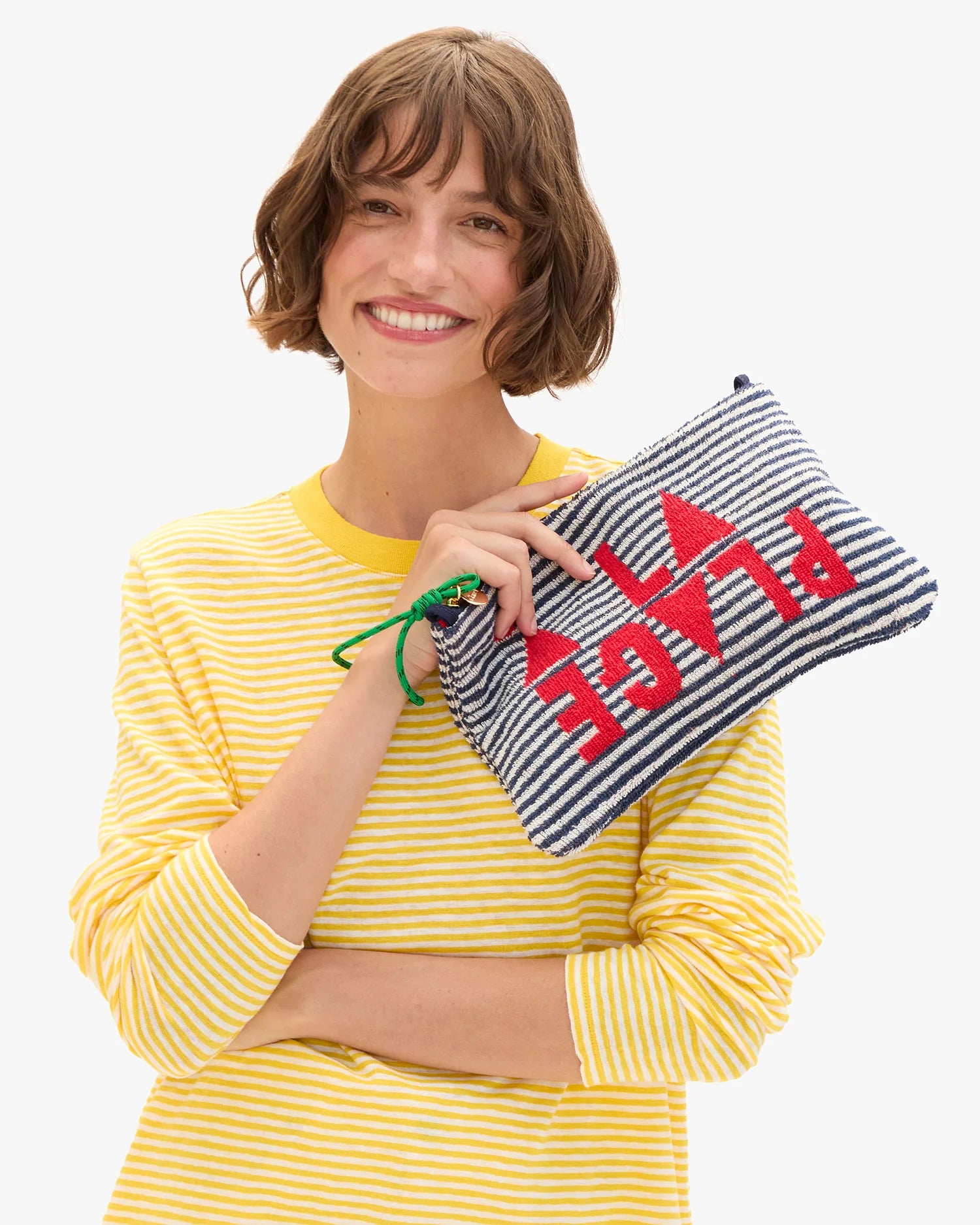 A woman with short brown hair in a yellow and white striped shirt smiles, holding the Clare Vivier La Plage Flat Clutch w/tabs, which features blue and white stripes and "PLACE" in red letters upside down.