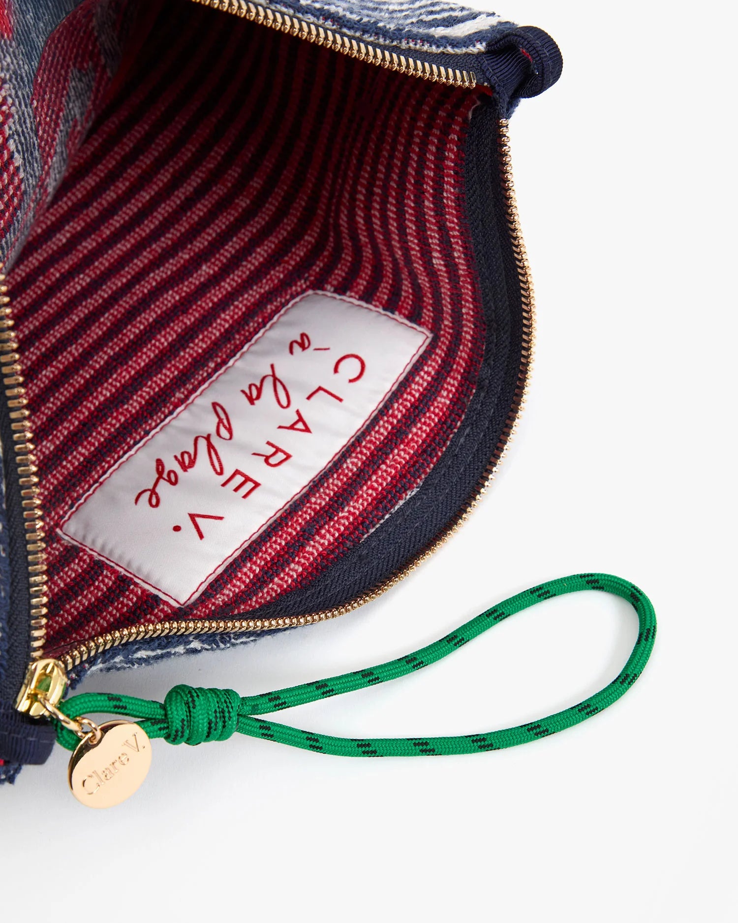 A close-up of the Clare Vivier La Plage Flat Clutch w/tabs shows its red striped lining, a white "Clare Vivier" label, and a green wristlet attached to the gold zipper pull.