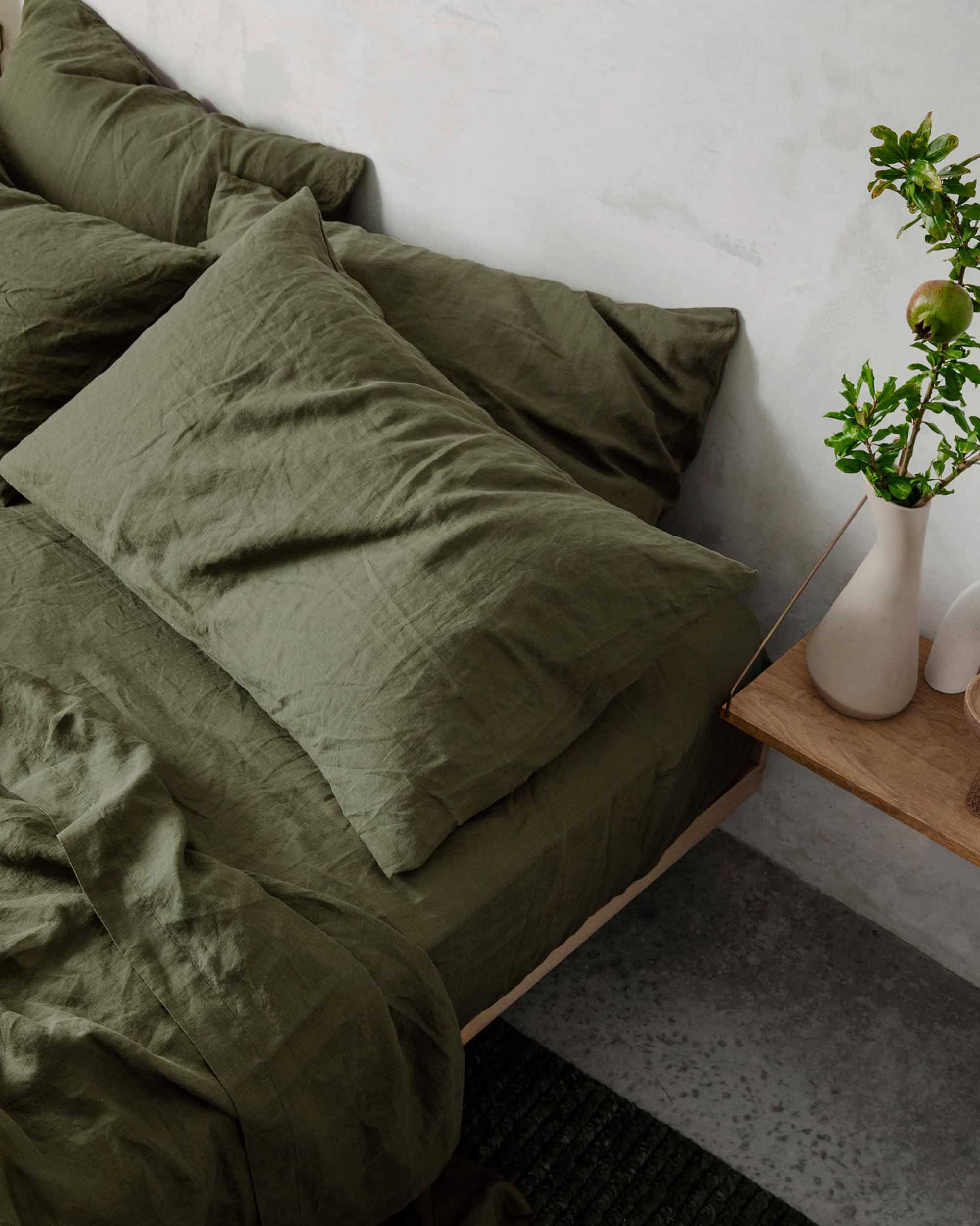 Olive green linen pillowcases by Cultiver Goods on a bed beside a wood nightstand with a white vase and greenery.