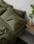 Olive green linen pillowcases by Cultiver Goods on a bed beside a wood nightstand with a white vase and greenery.