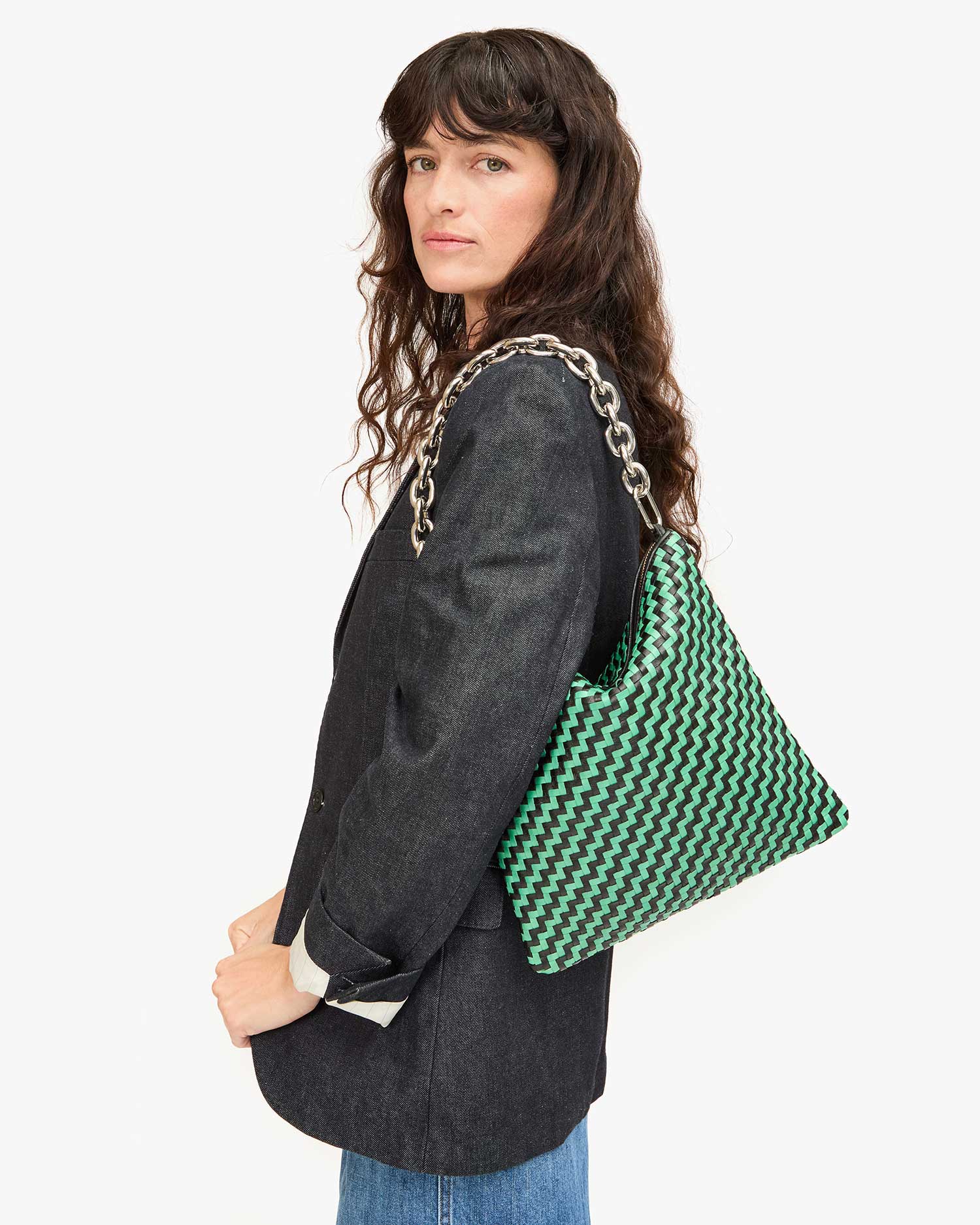 A person with long, wavy hair wearing a black blazer and jeans carries the Clare Vivier Foldover Clutch w/Tabs in green and black crocheted cotton over their shoulder. The background is plain white.