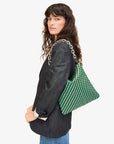 A person with long, wavy hair wearing a black blazer and jeans carries the Clare Vivier Foldover Clutch w/Tabs in green and black crocheted cotton over their shoulder. The background is plain white.